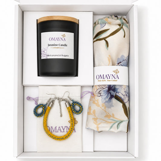 A curated gift set in a white box from OMAYNA, featuring a hand-poured jasmine candle in a black jar with a bamboo lid, a cream floral silk nscarf, and a small beaded pouch with matching beaded earrings.