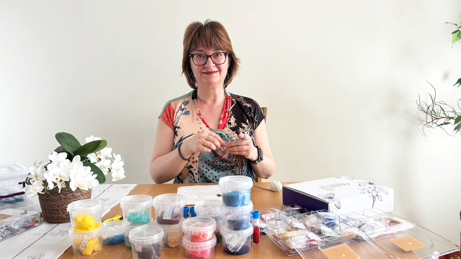 Theodora - the creator of Omayna, sitting at a table while making beaded jewellery, surrounded by containers with beads and plants