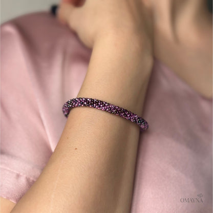 Close-up of a wrist wearing a purple beaded bracelet with a blurred pink fabric background.