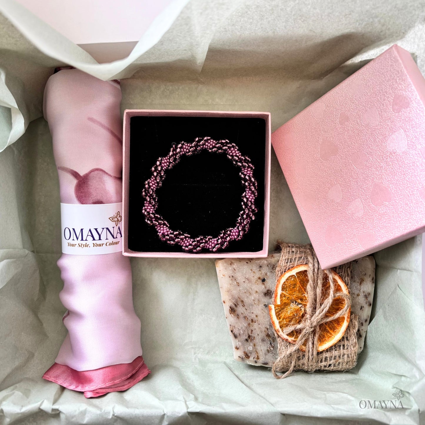 Gift box with pink scarf, bracelet, soap, and dried oranges on a light background