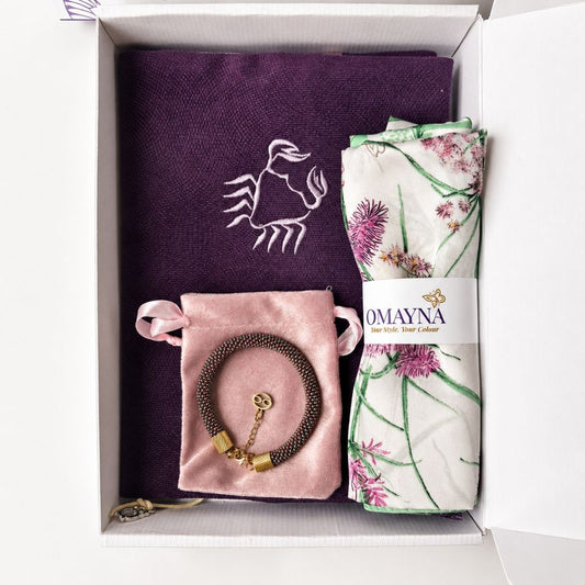 An open white Omayna gift box placed on green grass, containing a handmade bracelet, and a green patterned silk scarf wrapped in a branded paper band.