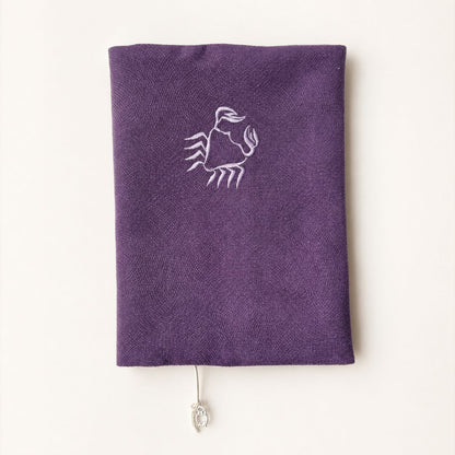 A purple textile book sleeve with a white embroidered Cacer sign symbol on the front, shown against a plain off-white background.