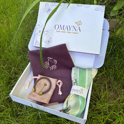 An open white Omayna gift box placed on green grass, containing a handmade bracelet, and a green patterned silk scarf wrapped in a branded paper band.