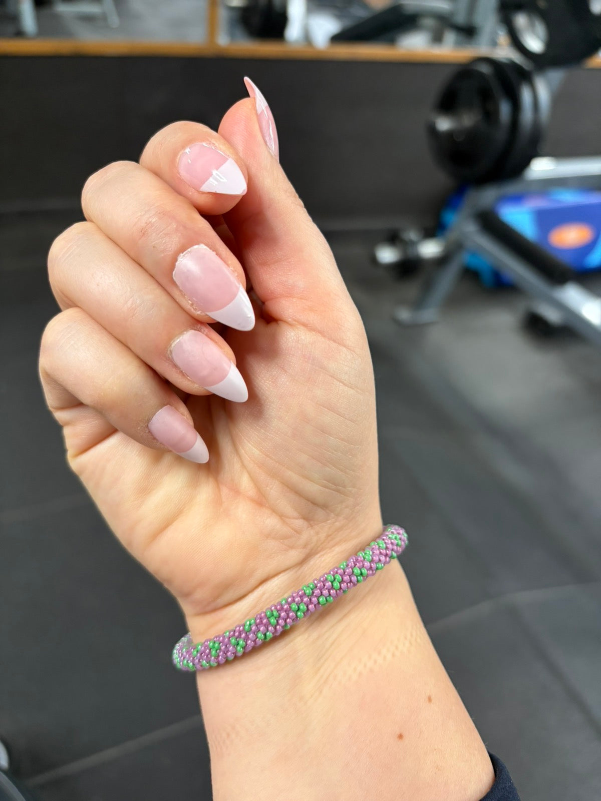 Hand with pink nail polish and a beaded bracelet in purple and green against a blurred indoor background - gym