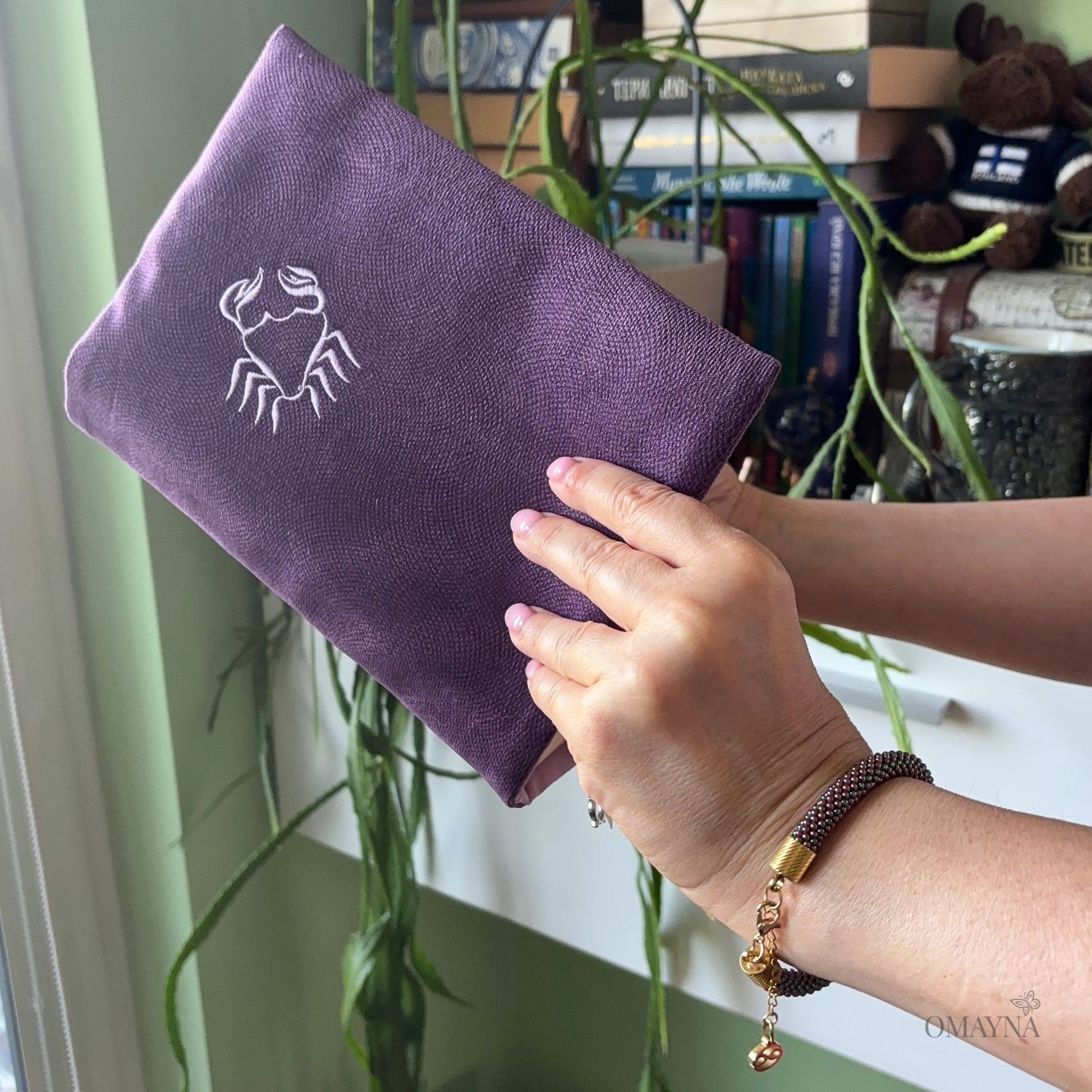 A person holds a purple book sleeve with a light purple Cancer symbol, set against a cozy indoor background with plants and book. A beaded bracelet is on a woman's hand.