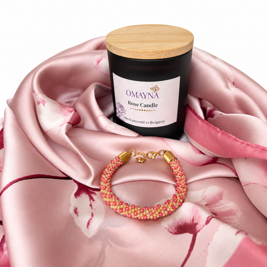 A black candle jar with a light wood lid and a white label reading OAYNA Rose Candle, styled on pink satin fabric with a pink and gold beaded bracelet in front.