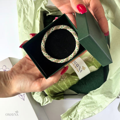 Hand holding a green jewelry box with a beaded bracelet, branded 'OMAYNA'.