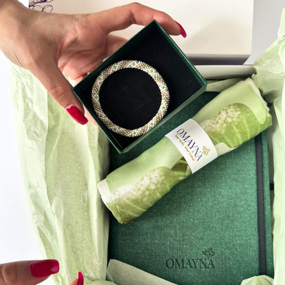 Hand holding a bracelet in a green box with a green silk scarf and Omayna branding.