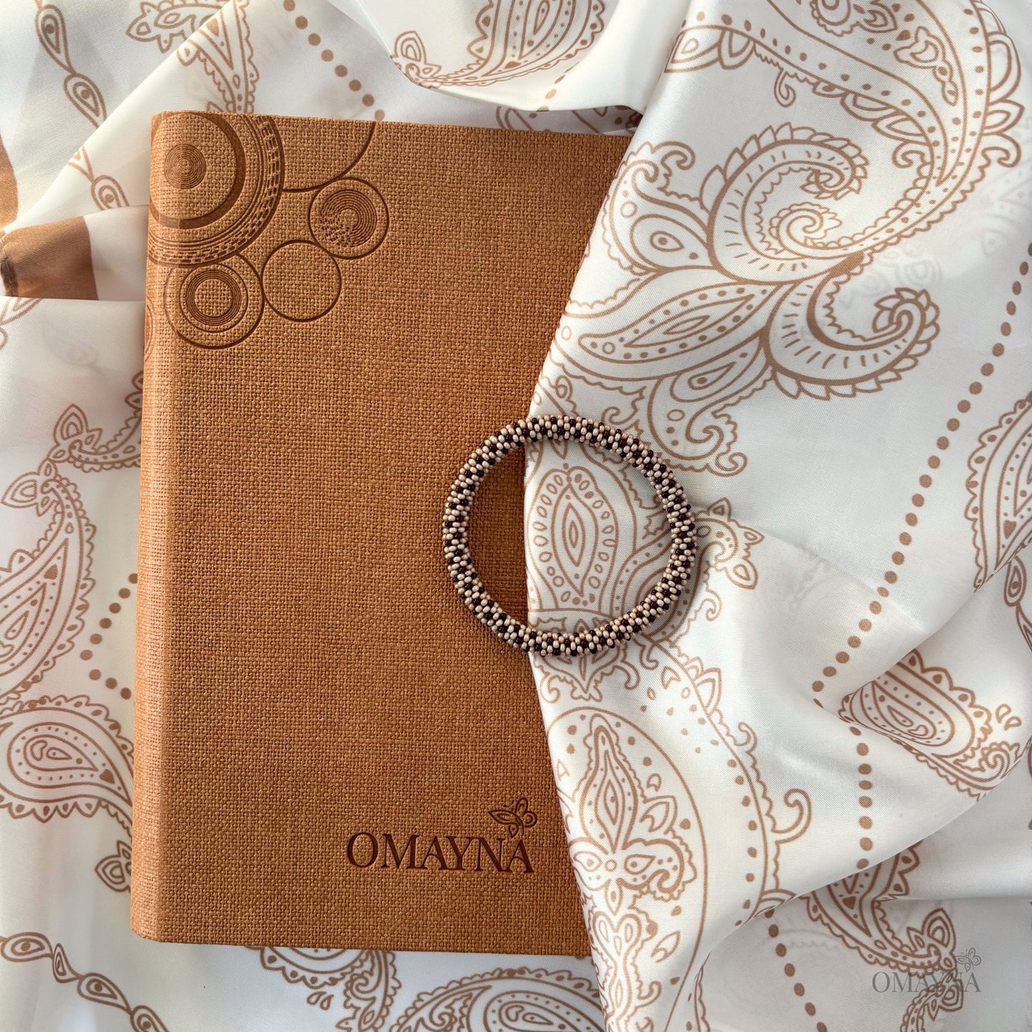 Beaded bracelet in beige and brown, Brown notebook with embossed design and 'OMAYNA' branding on a patterned silk scarf background