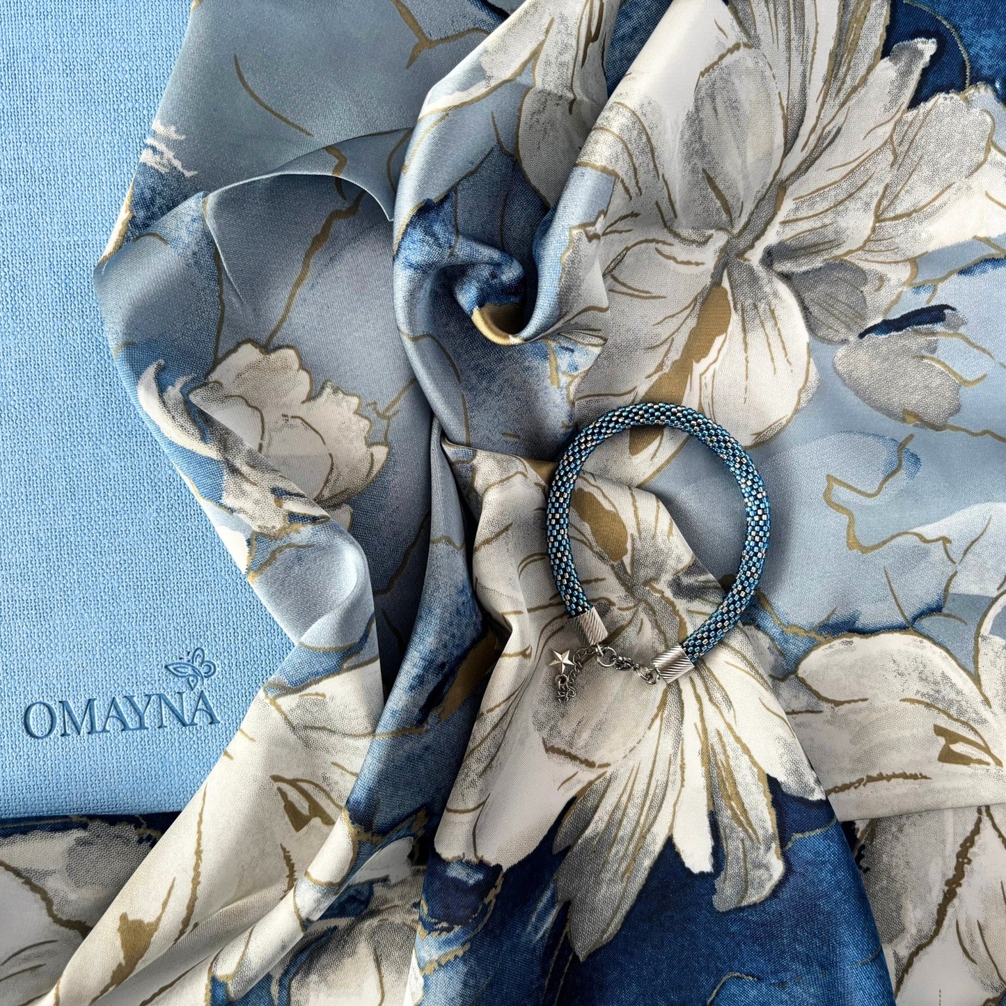 Floral-patterned scarf with a bracelet and notebook on a blue background, featuring the brand 'OMAYNA'.