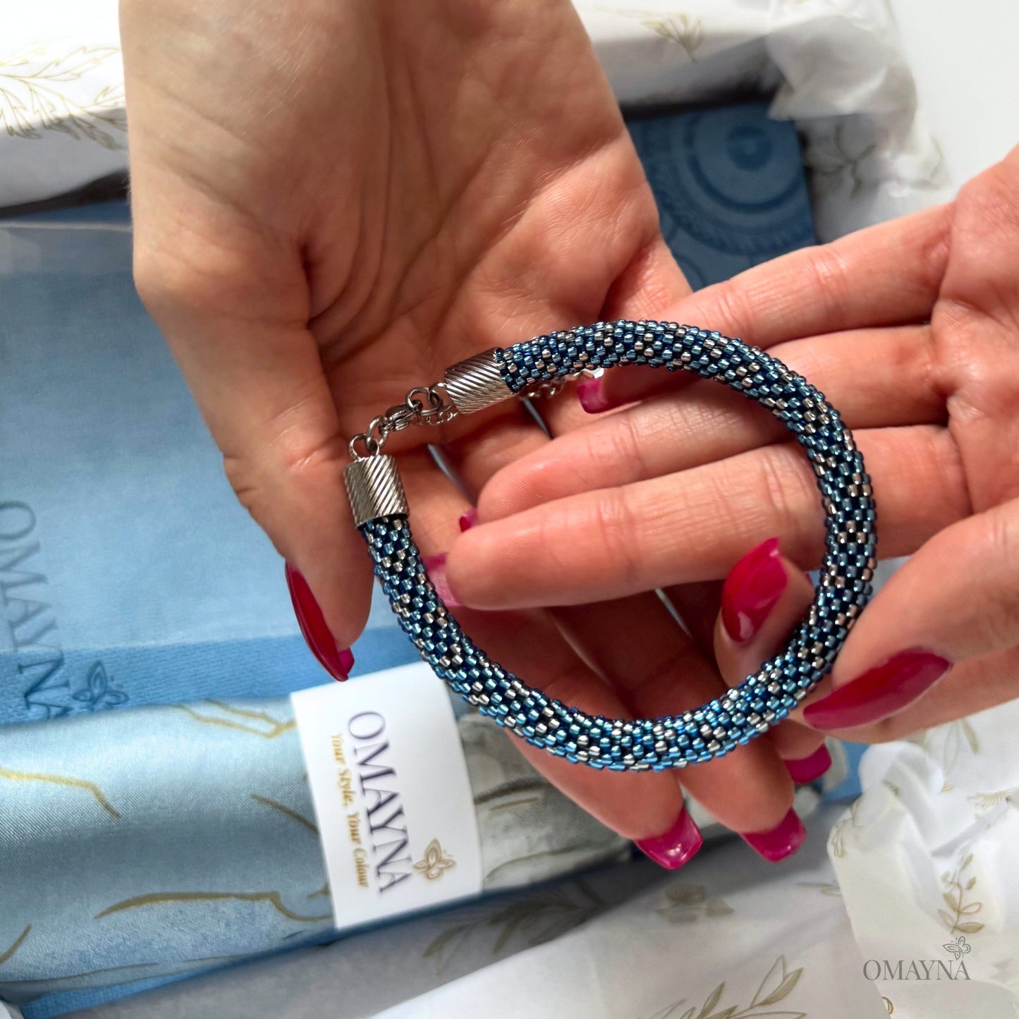 Hand holding a blue beaded bracelet with a visible brand tag Omayna in the background