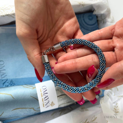 Hand holding a blue beaded bracelet with a visible brand tag Omayna in the background