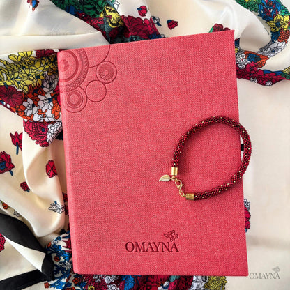 Red notebook with 'Omayna' branding and a bracelet on top, surrounded by a colourful scarf with a floral motif