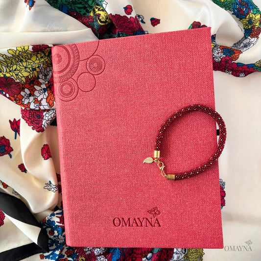Red notebook with 'Omayna' branding and a bracelet on top, surrounded by a colourful scarf with a floral motif
