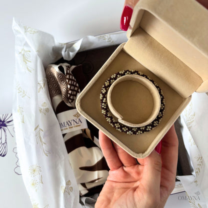 Beaded bracelet in dark brown and pearl in a box held by a hand with Omayna branded background